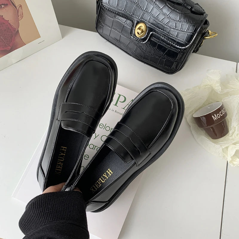 Black loafers with 'HUGO' branding on a white surface with a black handbag and coffee cup in the background.