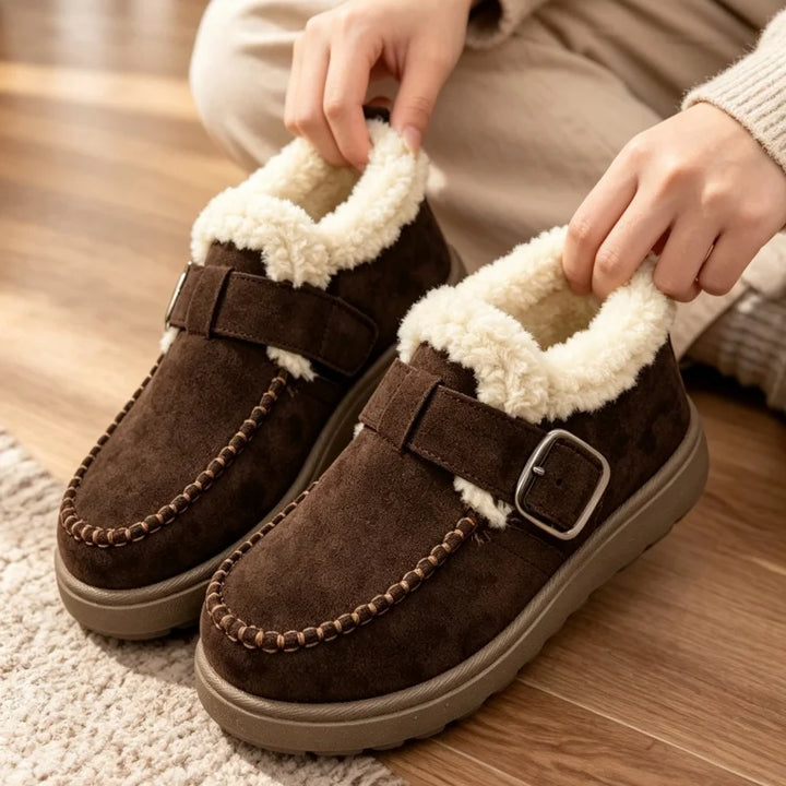 A pair of women's brown suede slip-on shoes with cream sherpa lining and decorative stitching, shown on a wooden floor with a person's hands adjusting them.