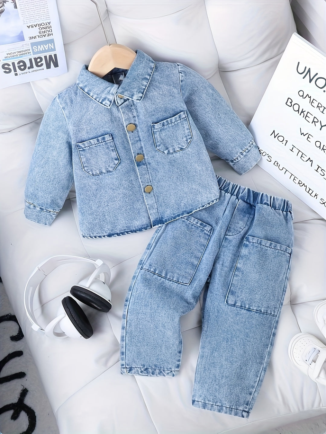 Children's denim jacket and pants set on a white surface with a newspaper and sign in the background.