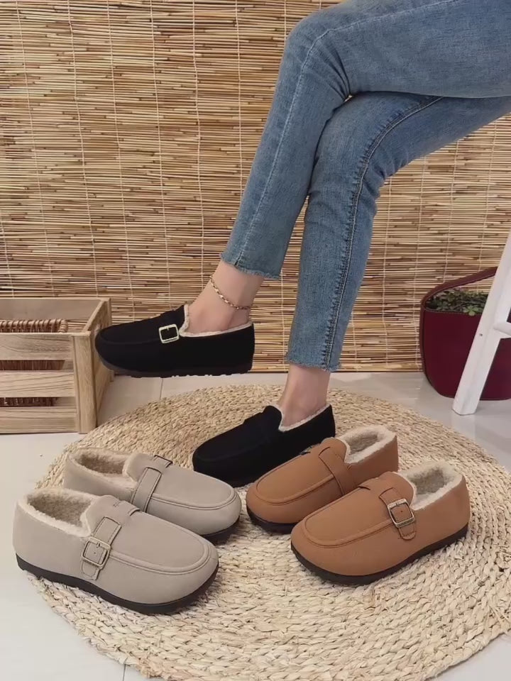 Video of women's slip-on mule loafers with soft shearling lining and buckle straps are displayed on a woven mat. One pair is black, one is light beige, and one is tan/caramel.