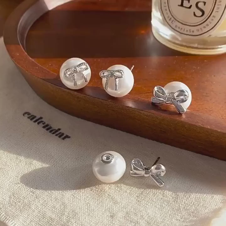Hand wearing a decorative earring with a bow design on a neutral background